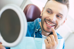A happy man sitting in a dentist office, smiling and admiring his new dental implant in a hand -held mirror.